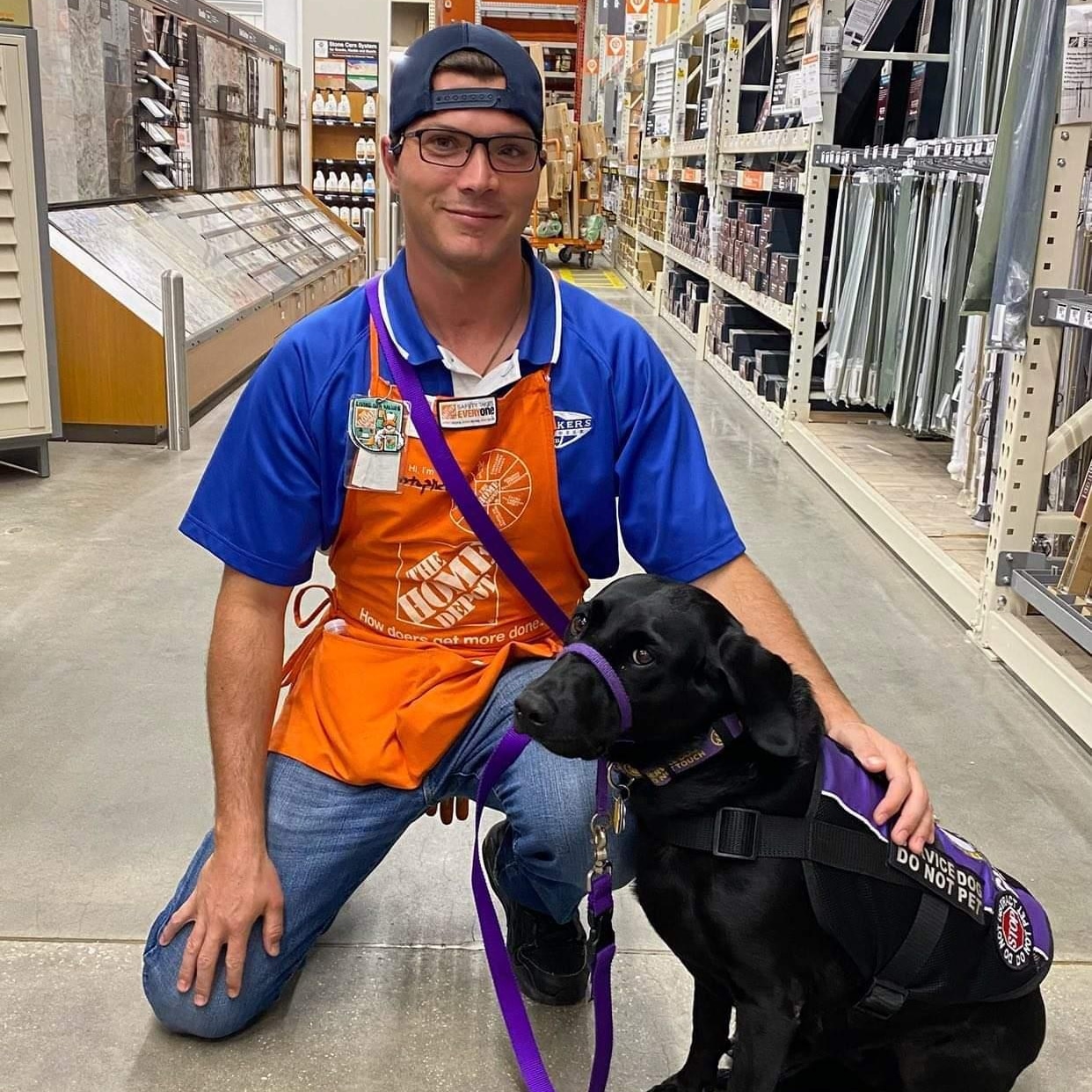 Man, wearing Home Depot apron in the store with black service dog, wearing a vest sitting next to him